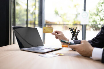 A man is using a laptop and a cell phone to pay for something. He is holding a credit card and a debit card
