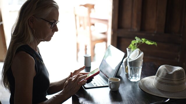 A mature woman sits at a cafe table engrossed in her tablet and phone while enjoying a coffee. She embodies the digital nomad lifestyle in a warm environment.