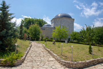 historic stone building with domed roofs surrounded by lush greenery, clear blue sky above, paved...