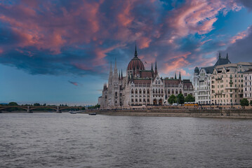 Obraz premium The Hungarian Parliament Building in Budapest stands by the Danube River under a colorful sunset sky, with a bridge and historic buildings in view.