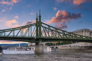 Fototapeta premium The Liberty Bridge spans the Danube River in Budapest, Hungary, under a vibrant sunset sky. Riverboats and historic buildings line the riverbank below.
