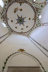 An upward view of an ornate Islamic dome featuring a central star design, decorative green and gold leaf patterns along the arch below, and a rectangular tomb marker at the bottom of the frame.
