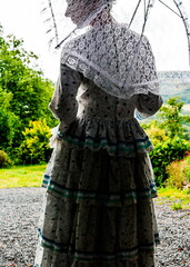A person in a vintage dress and lace head covering stands under a clear umbrella, looking out at a...