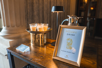 A wooden table displays a welcome drink sign, a metal pitcher, and glasses. A black lamp and warm wooden tones create a cozy, elegant atmosphere.