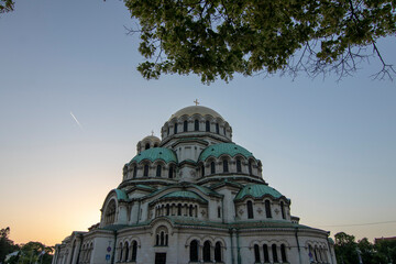 Fototapeta premium Alexander Nevsky Cathedral in Sofia framed by tree branches at sunset, showcasing its majestic Neo-Byzantine architecture and iconic green domes against a clear evening sky.