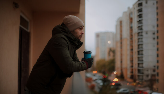 Young man in winter clothes holding a coffee cup, leaning on a balcony railing, gazing thoughtfully at a cityscape with tall buildings during a cold, overcast evening.