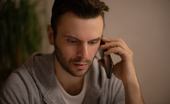 Fototapeta Young man in casual hoodie having a serious phone conversation indoors, focused and concerned, in a dimly lit home setting with warm tones and a blurred plant in the background.