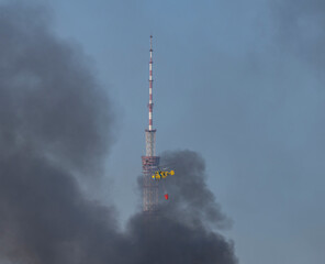 A yellow firefighting helicopter drops water over heavy smoke after a missile strike in Kyiv, with a TV tower in the background during the Russia-Ukraine war.