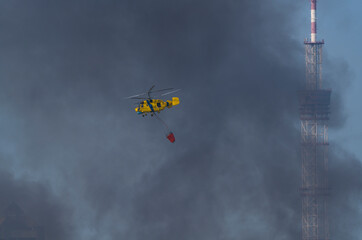 A yellow firefighting helicopter drops water over heavy smoke after a missile strike in Kyiv, with a TV tower in the background during the Russia-Ukraine war.