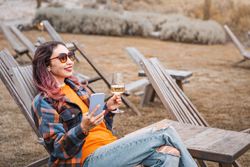 Tourist enjoying a glass of wine while using her smartphone, sitting on a wooden deck chair in a winery garden