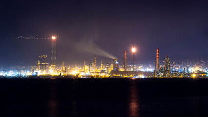Fototapeta premium Nighttime panoramic view of an industrial oil refinery by the sea, with smoke, flares, and glowing chimneys, illustrating energy production, pollution, and environmental impact.