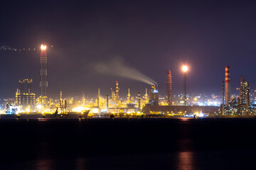 Fototapeta premium Brightly lit oil refinery at night with flare stacks, smoke emissions, and glowing chimneys by the sea, reflecting industrial energy production and environmental pollution issues.