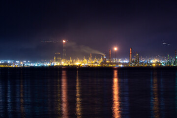 Nighttime oil refinery glowing with industrial lights, smoke stacks emitting vapor, and flaring towers reflecting vividly on calm water, showcasing continuous energy production and heavy industry.