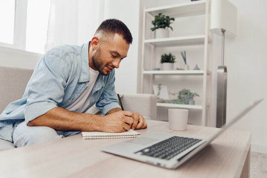Smiling man engaged in work, writing notes with a laptop nearby, showcasing a cozy home office environment filled with greenery and modern decor.