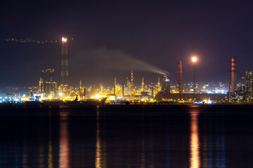 Fototapeta premium Oil refinery at night glowing with bright lights, smoke stacks, and flaring towers reflecting on water, capturing the power, energy, and industrial strength of modern oil production.