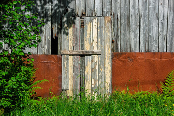A weathered barn features a rustic wooden door, partially open, with vibrant green grass and wildflowers nearby under clear blue skies.
