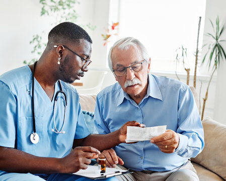 Doctor or nurse caregiver showing a prescrption or documents and holding drug bottle to senior man at home or nursing home
