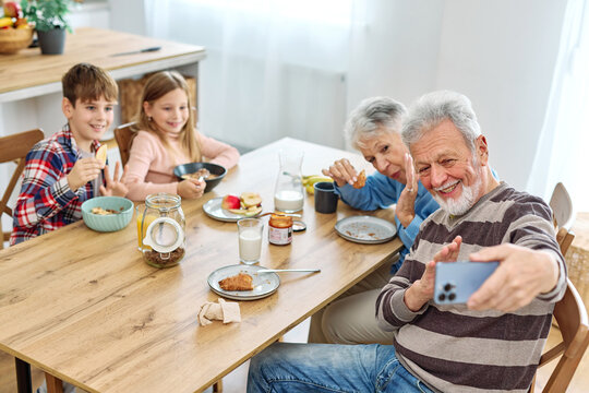 Portret of brother and sister, grandchildren and grandparents having fun together taking a selfie photo with a mobile phone and eating breakfast in kitchen