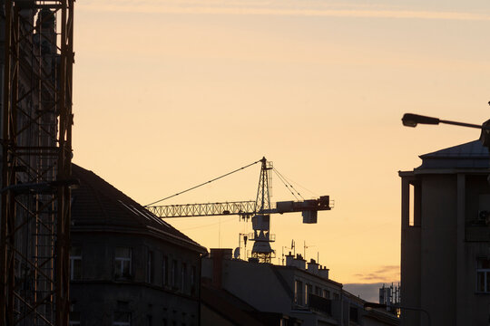 A striking silhouette of a construction crane stands boldly against the vivid colors of a stunning sunset sky, surrounded by an array of urban buildings that define the city - Powered by Adobe
