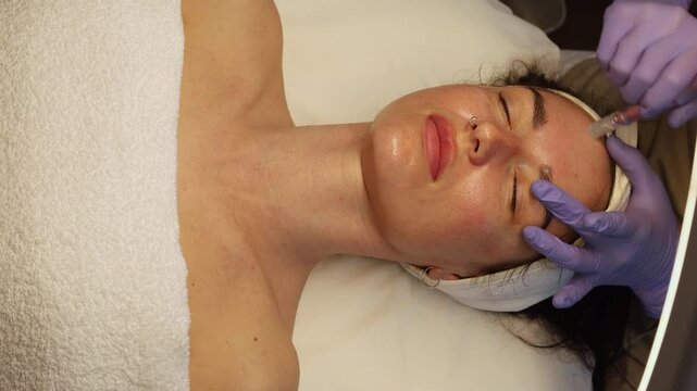 Woman receiving a professional microneedling treatment at a cosmetology clinic. A beautician in gloves is using a dermapen device on the client's face for skin rejuvenation and collagen stimulation