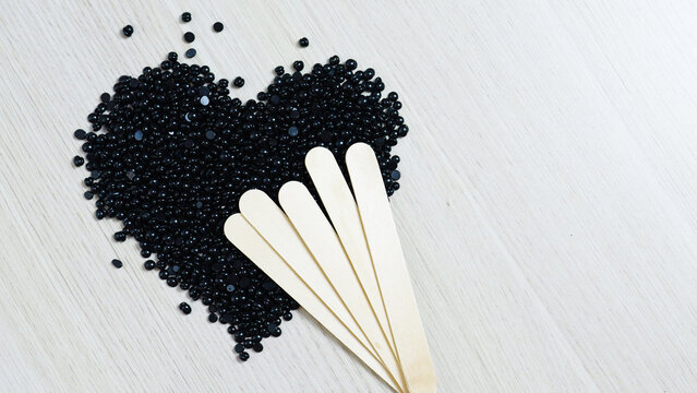 Black hard wax beans forming heart shape on rustic wooden surface, wooden spatulas nearby, representing beauty, hair removal, and skincare preparation