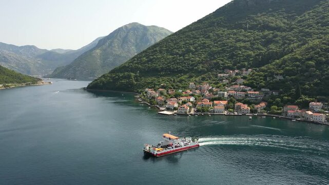 A passenger ferry sails along the Verige Strait in the Bay of Kotor against the backdrop of a picturesque mountain coastline on a sunny summer day, aerial view