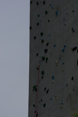 A textured climbing wall with colorful holds and a safety rope extends upward under a muted sky, conveying challenge and vertical adventure