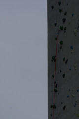 A textured climbing wall with colorful holds and a safety rope extends upward under a muted sky, conveying challenge and vertical adventure