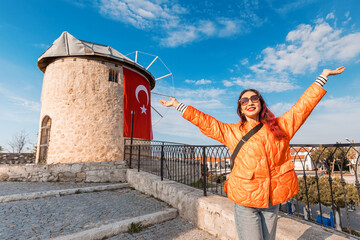 Woman joyfully raising arms near a historic stone windmill with a Turkish flag, under a bright blue sky, symbolizing freedom and happiness