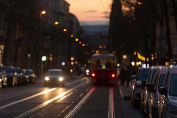 As the sun sets and twilight envelops the city, a colorful tram glides along a bustling street, surrounded by parked cars and soft glowing lights enhancing the vibrant atmosphere