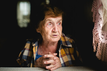 An elderly woman sits quietly, hands folded, gazing thoughtfully toward the window. Her face is...