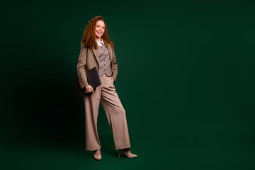 Confident young businesswoman with red curly hair in formalwear posing against an emerald green...