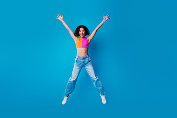 Joyful young woman jumping enthusiastically against a vibrant blue background in fashionable casual...