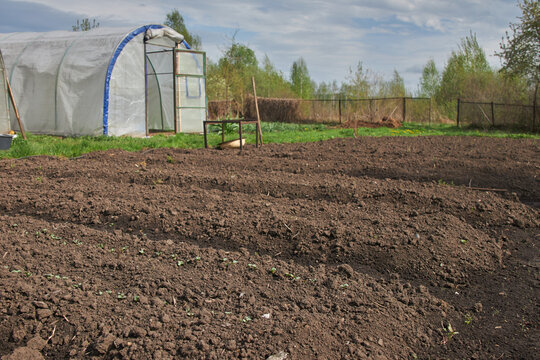 A freshly tilled garden plot awaits planting beside a greenhouse under a cloudy sky.