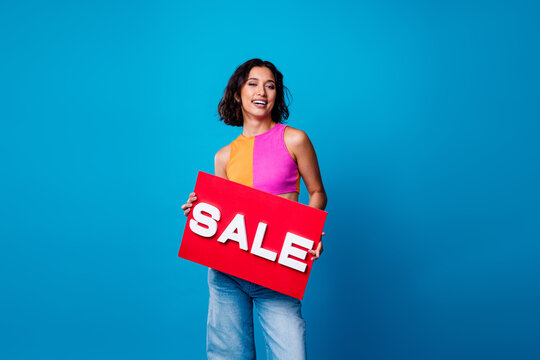 Young woman with short brunette hair holding a red sale sign, smiling against bright blue background, stylish outfit