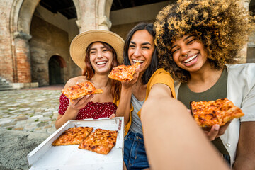 Three multiracial women eating pizza in city street - Smiling female friends taking selfie enjoying summer vacation in Italy