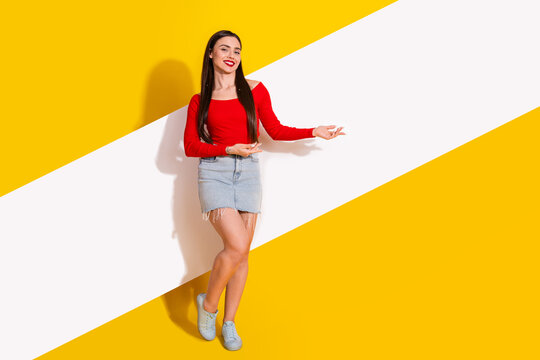 Fototapeta Smiling young woman in a red shirt and denim shorts posing against a bright yellow geometric background showcasing style and fashion.