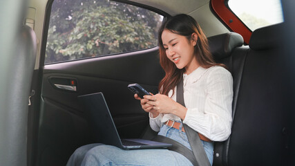 A smiling young woman is sitting in a car with a laptop on her lap while looking at her mobile...