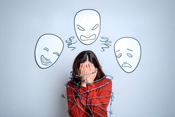 A person in a red shirt, entangled in barbed wire and covering their face, stands before three cartoon masks on a wall—laughing, angry, and sad—symbolizing emotional conflict.