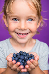 Adorable kid holding blueberry fruit and smiling over violet studio background. Healthy relationship with food and child nutrition concept. Healthy baby food with vitamins and kids vegetarianism