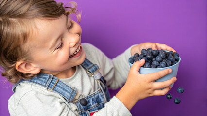 Adorable kid holding blueberry fruit and smiling over violet studio background. Healthy relationship with food and child nutrition concept. Healthy baby food with vitamins and kids vegetarianism