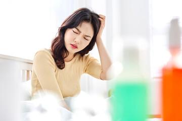 Naklejka premium Young Asian Woman in Yellow Sweater Sitting on Bed, Holding Head in Pain, Suffering from Headache in Bright Morning Light