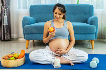 Asian pregnant woman sits on yoga mat on living room floor, hold apple after exercises at home. Beautiful pregnancy mother with big belly eats healthy food and do workout for good health. Happy family