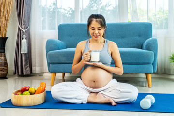Eight months pregnant Asian woman wearing a sports bra sitting on a sofa stroking her belly and drinking calcium-fortified milk at living room at home, pregnancy and motherhood concept.
