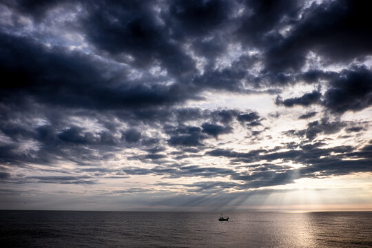 A lone boat on a vast ocean under a dramatic cloudy sky with rays of sunlight breaking through