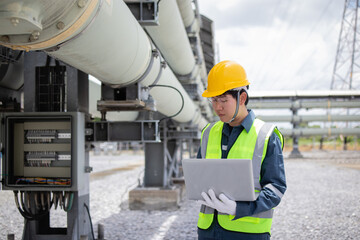 Engineer is working at power station inspecting large industrial pipes while using laptop to ensure efficient operation and safety in outdoor facility environment