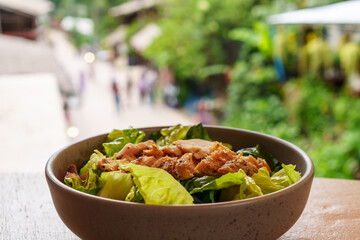 Spiced Chicken Salad on wooden table with blurry Mae Kampong village street background, small village with traditional houses and beautiful nature. Destination of traveler. Chiang Mai, Thailand.