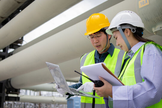 Engineer working at power station wearing safety helmet and reflective vest while checking laptop and clipboard with walkie talkie in industrial environment - Powered by Adobe
