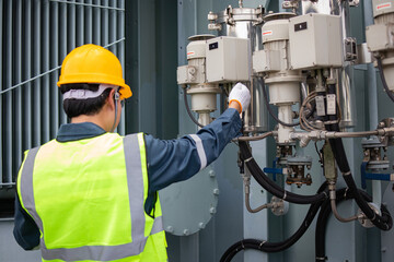 Worker wearing yellow helmet and reflective vest inspects oil filter attached to transformer inside...