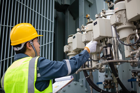 Technician wearing yellow helmet and safety vest inspects oil filter system at substation near large transformer ensuring proper maintenance and safety